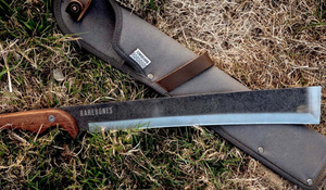 A large fixed-blade machete-style knife laid on grass, partially resting on a fabric sheath.