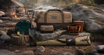 Field-tested outdoor gear kit with canvas duffel bag, wool blanket, shovel, leather cases, and compass by a riverside campsite