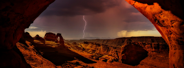 Lightning strikes beyond Delicate Arch in Utah, framed by a sandstone cave and dramatic storm clouds over the red rock desert. 